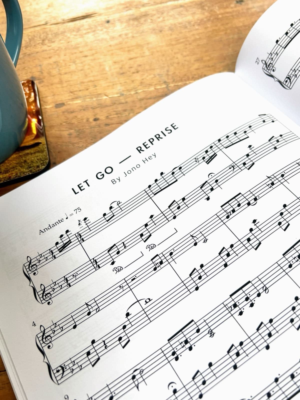 Printed sheet music for Let Go (Reprise) by Jono Hey, open on a wooden table beside a mug